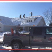 Three men clearing snow from a home’s roof with a truck parked in front.